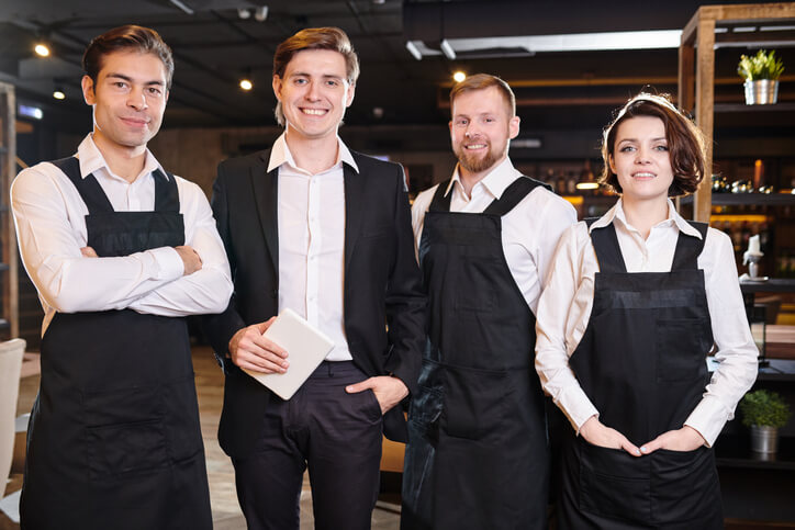 Three restaurant staff in black aprons stand confidently beside a manager in a suit holding a tablet. They pose as a united front, ready to provide excellent service in a modern, welcoming dining environment.